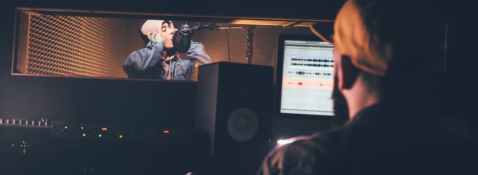 Recording session in a studio with a male artist at a microphone and sound engineer at a mixing desk.